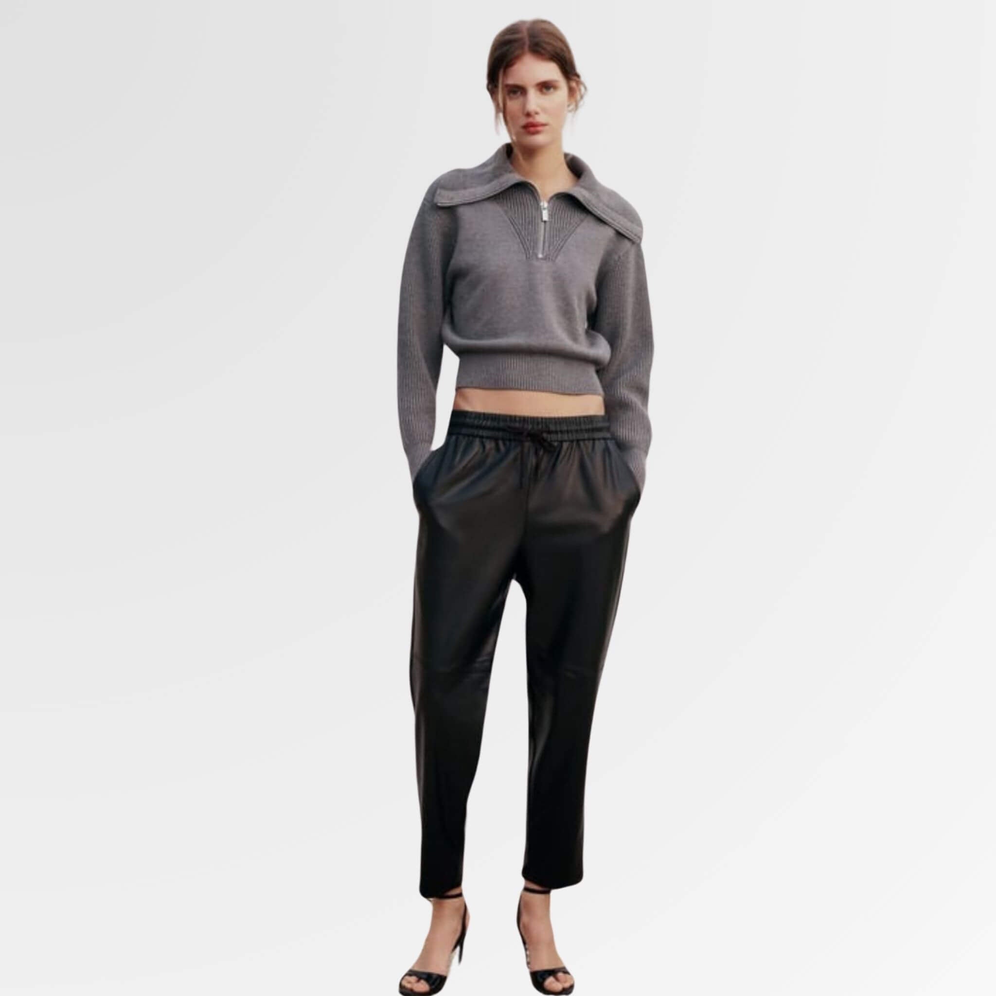 A stylish woman stands confidently, wearing high-quality leather pants, a half-zip sweater, and chic heels.