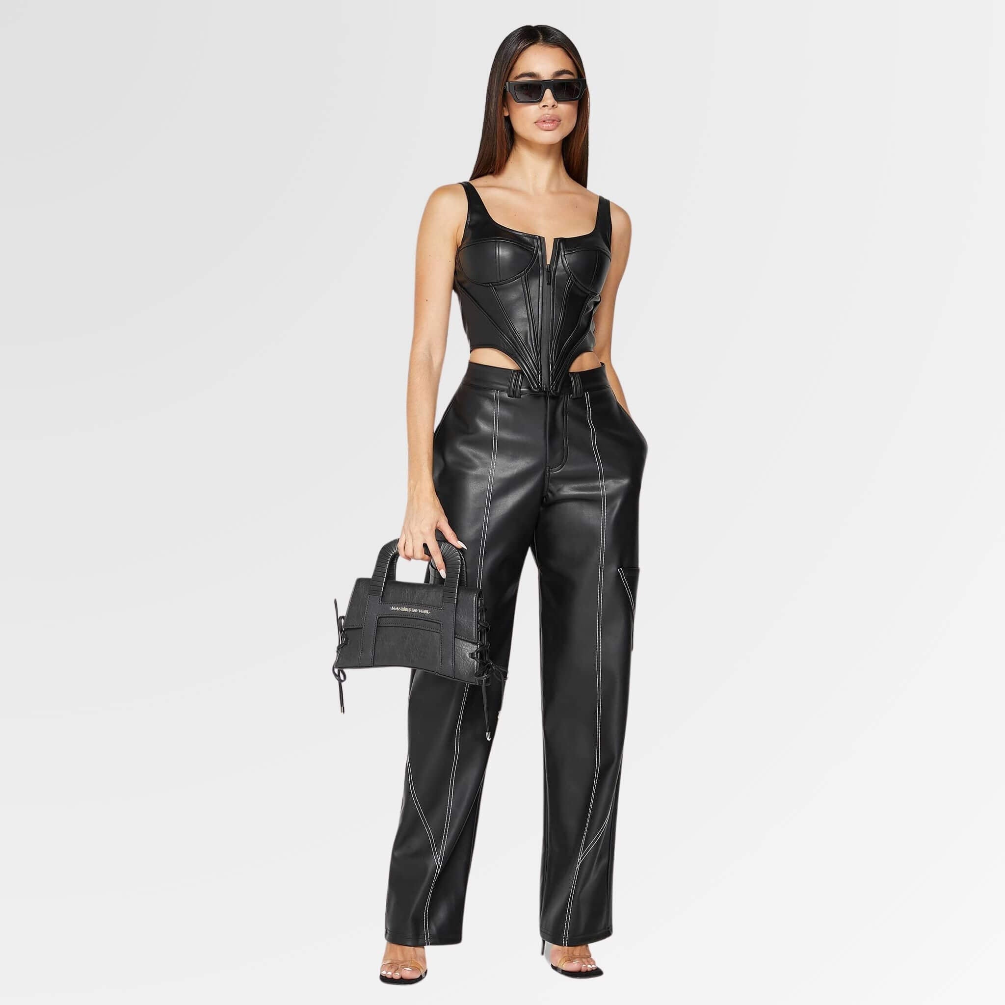 A stunning woman stands confidently, wearing a stylish leather corset top paired with sleek leather pants, accessorized with a chic leather bag and trendy black glasses.