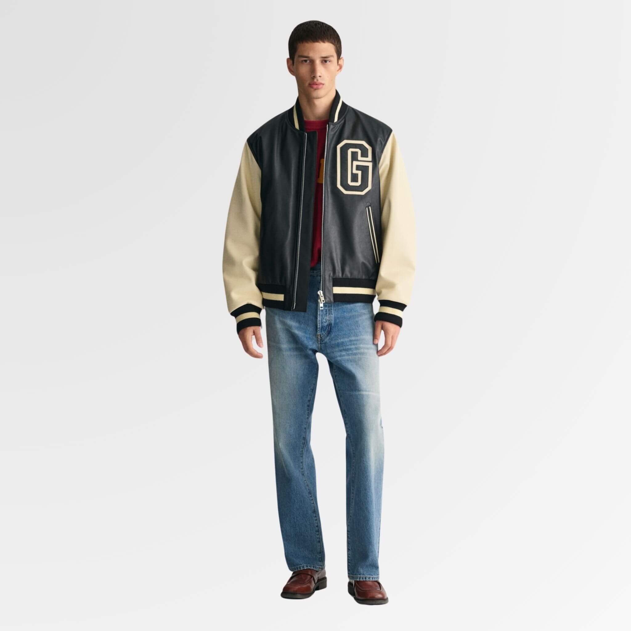 A young man standing tall in a leather varsity jacket and blue straight-fit jeans, looking directly at the camera with a confident expression.