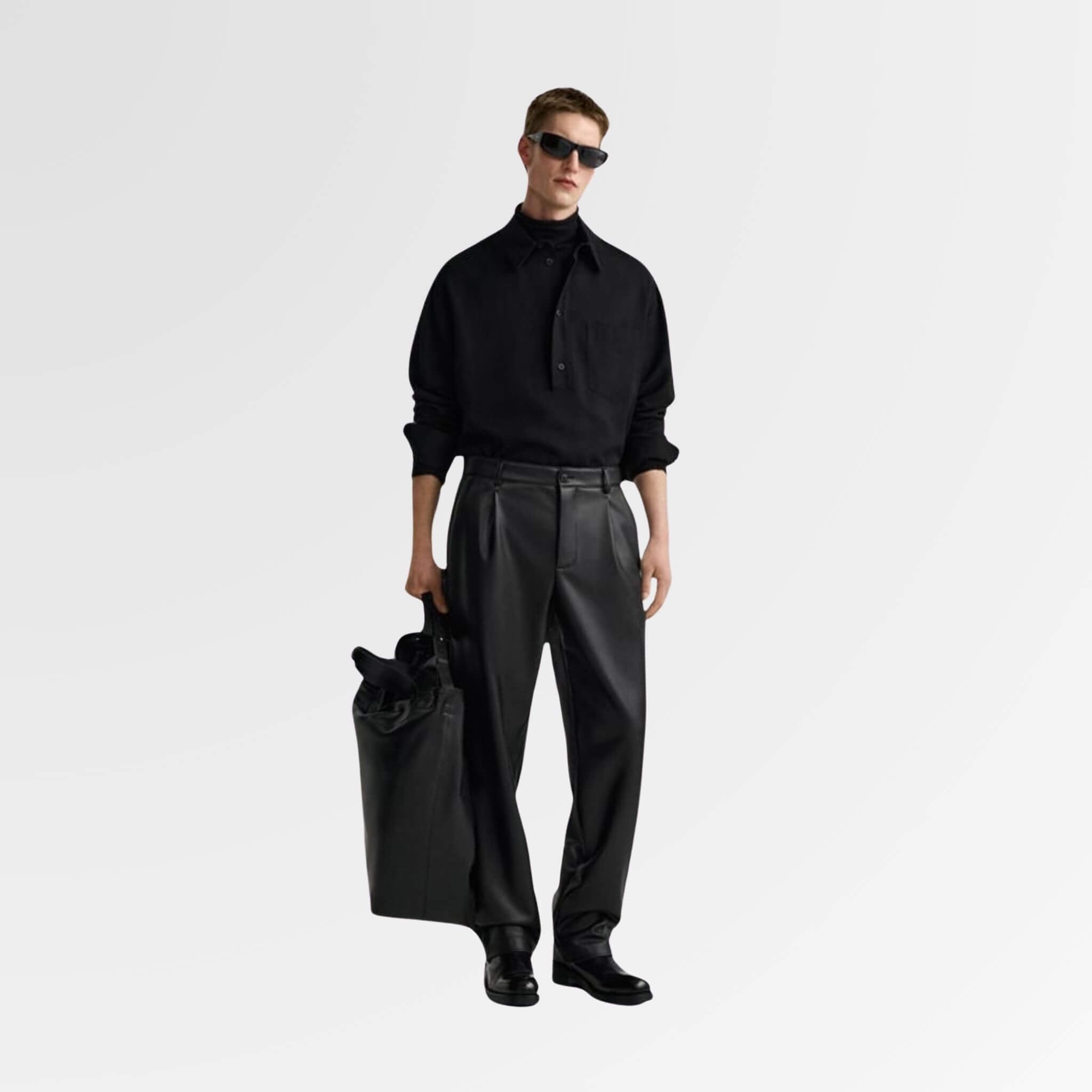A young man wearing a black button-down shirt and straight-fit leather pants, holding a bag in one hand, posing confidently with sunglasses on, showing full swag.