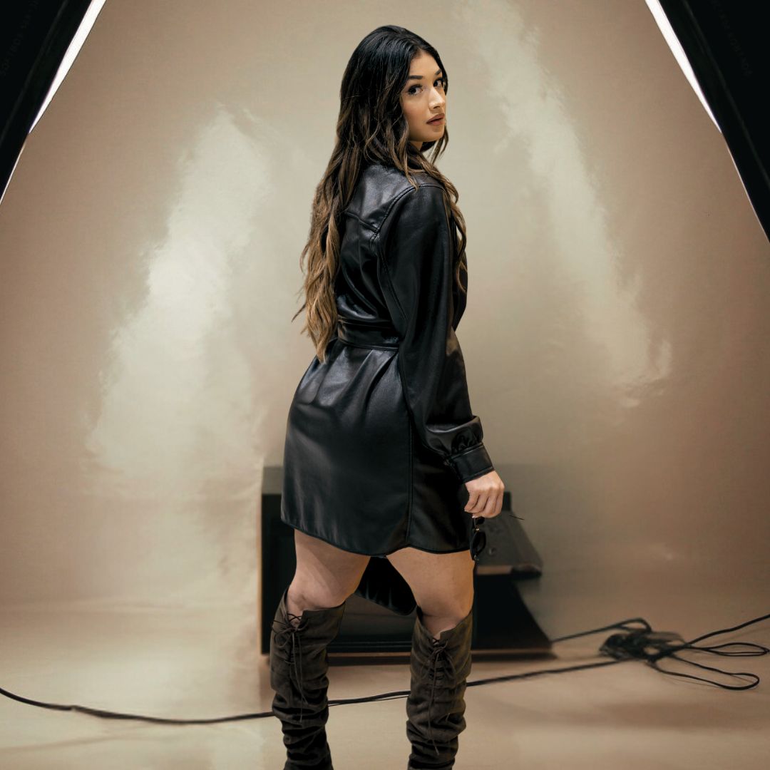 Woman in a black leather long jacket and knee-high boots standing in a studio setting.